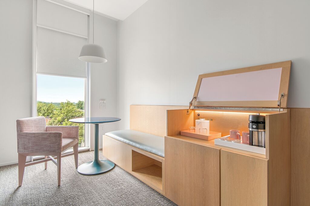 A modern hotel room with a wooden cabinet, a small table, a bench, and a large window offering a view of greenery.