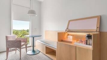 A modern hotel room with a wooden cabinet, a small table, a bench, and a large window offering a view of greenery.