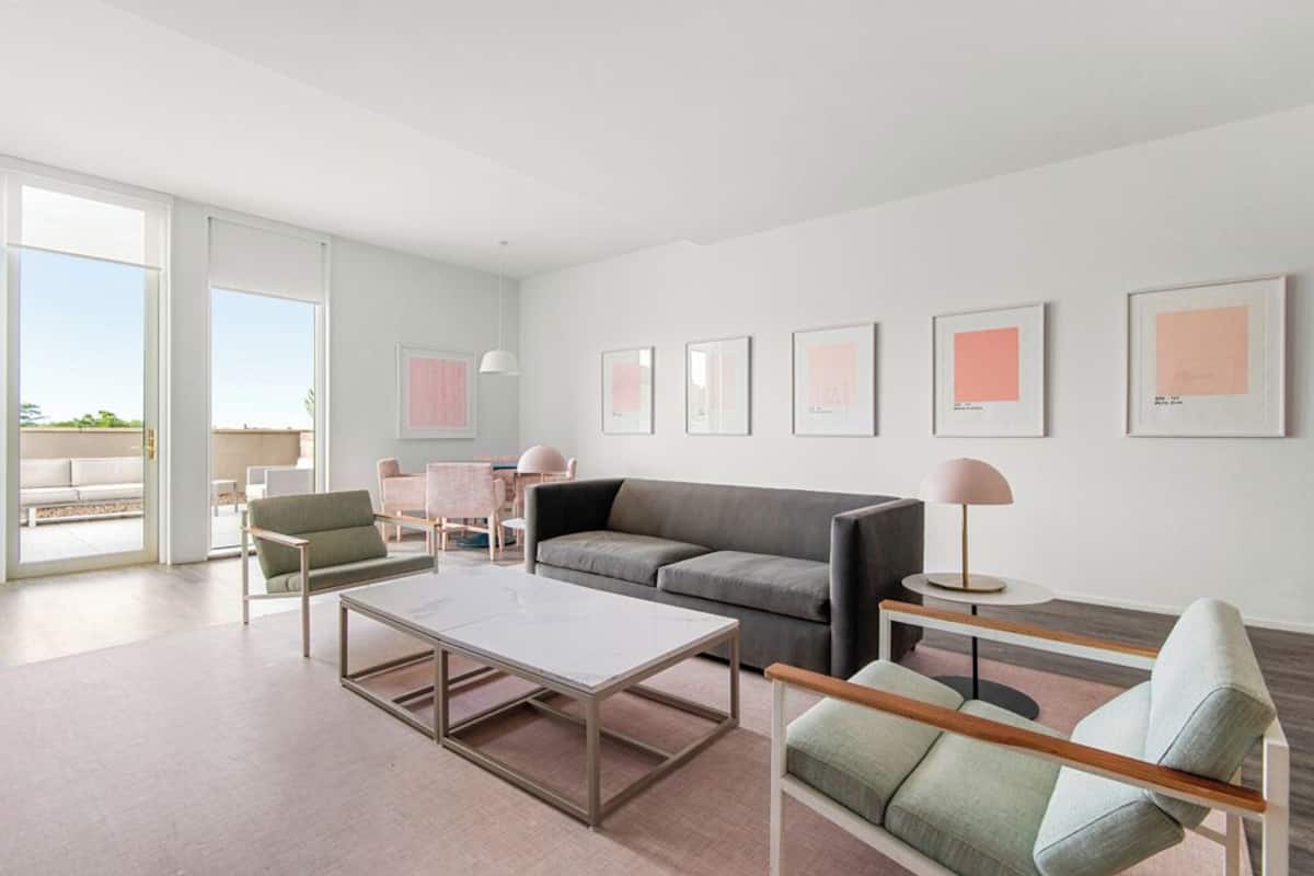 Una sala de estar moderna con un sofá, sillones, una mesa de centro y una lámpara. Hay cuadros enmarcados en la pared y un ventanal que proporciona luz natural.