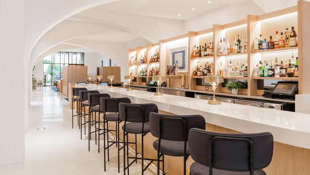 Een moderne bar met een lange toonbank, meerdere planken vol drankflessen en barstoelen.
