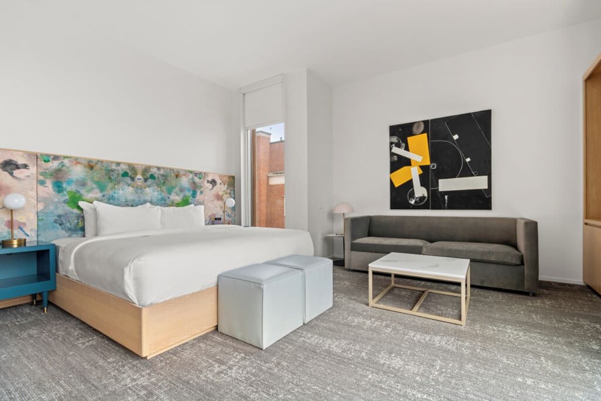 Una habitación de hotel moderna con una cama grande, un sofá gris, una mesita pequeña y arte abstracto en las paredes.