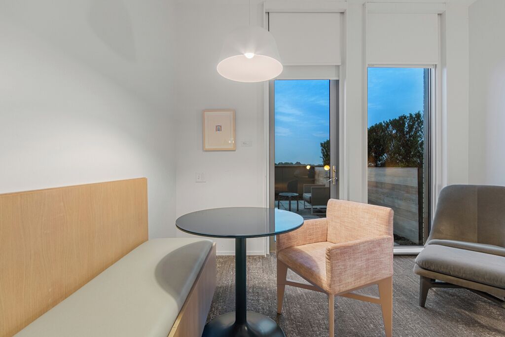 A modern hotel room with a round glass table, two upholstered chairs, a sofa, and a large window offering a view of the outdoors.