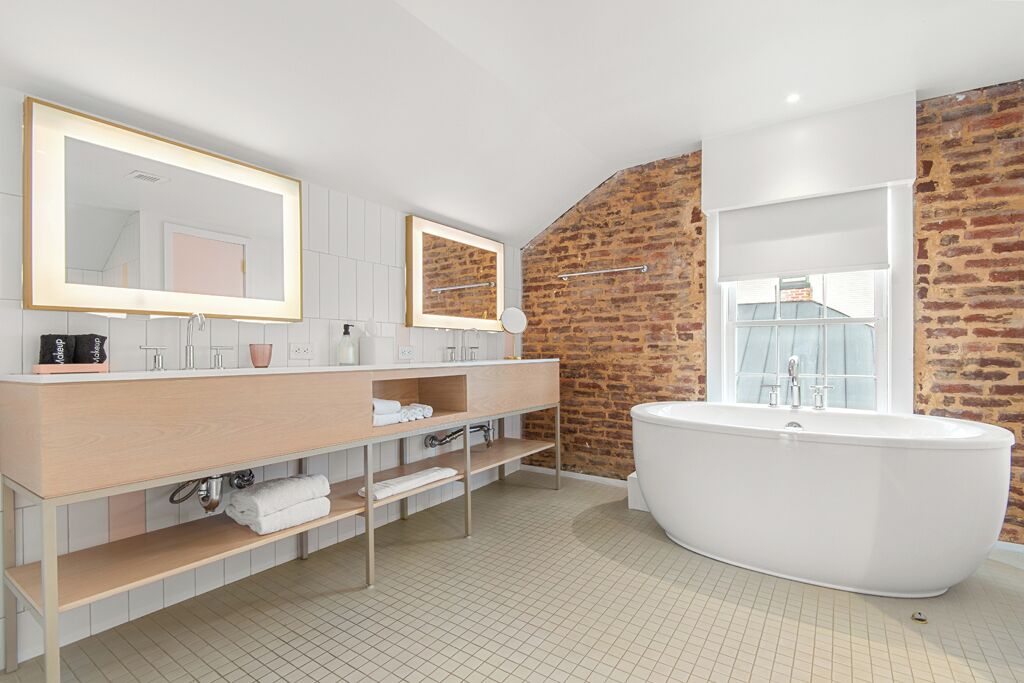 Un baño moderno con una bañera independiente grande, un tocador doble con espejos y una pared de ladrillo decorativa.