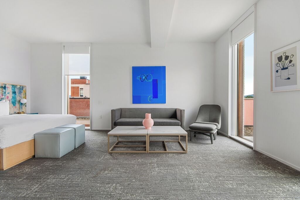 A modern hotel room with a bed, a gray sofa, a gray armchair, a coffee table, and a large window offering a view of the city.