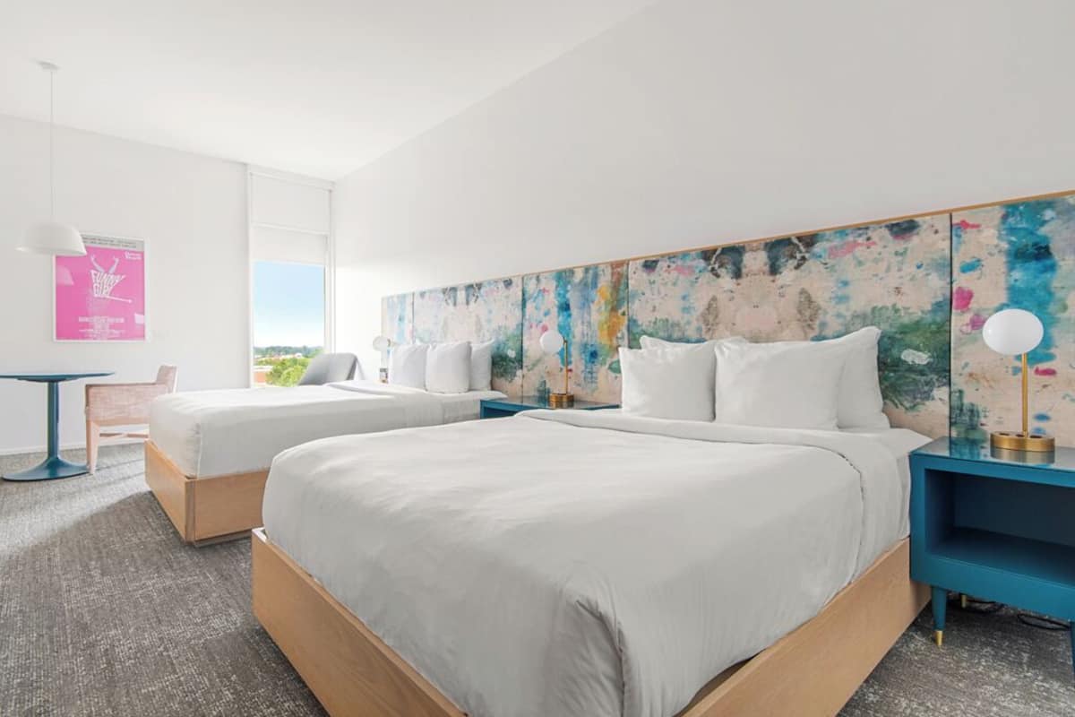 Habitación de hotel con dos camas, un cuadro abstracto grande en la pared, una mesita y vistas al exterior.