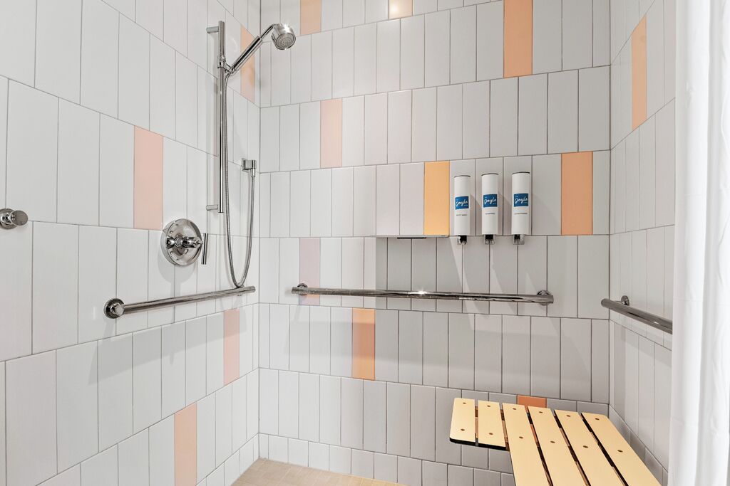 Suihkutilassa on lasiseinä, käsisuihku ja hylly, jossa on hygieniatuotteita.