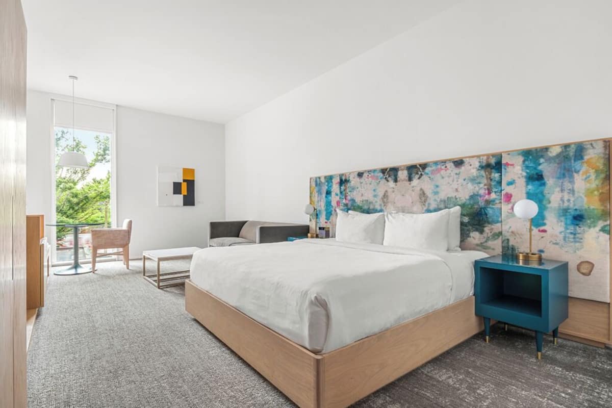 Un dormitorio moderno con una cama grande, un escritorio y una silla. Se ve follaje verde a través de la ventana.