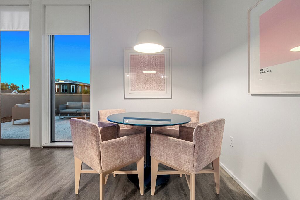Uma sala de jantar moderna com uma mesa redonda e quatro cadeiras, uma luminária pendente e uma obra de arte emoldurada na parede.