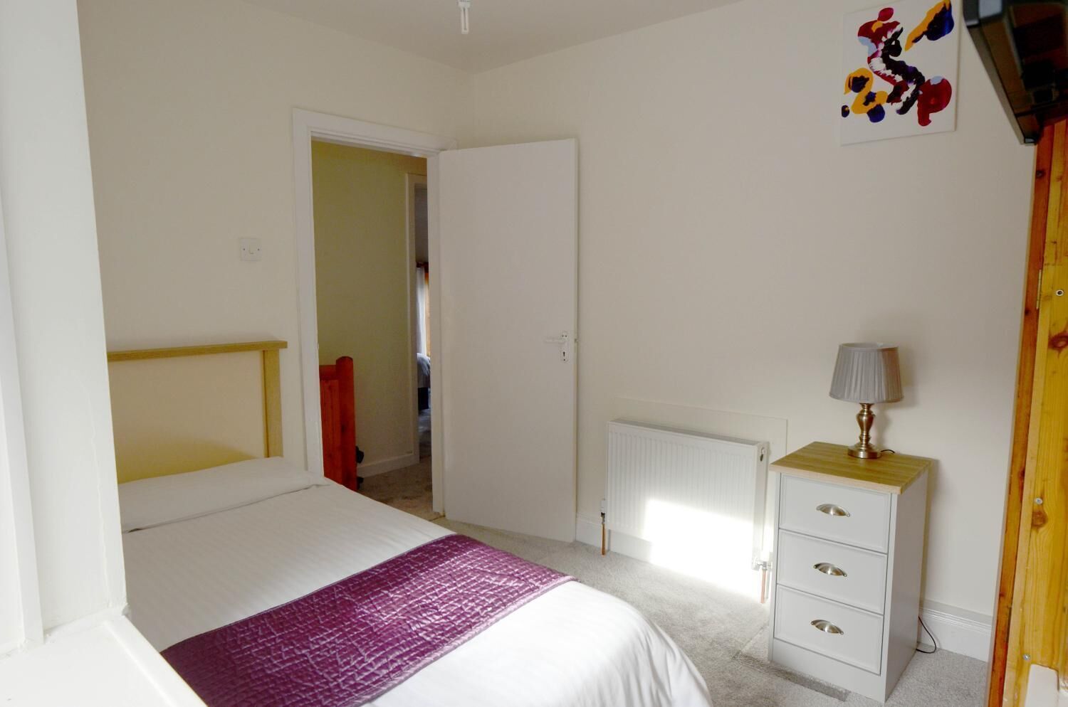 Une chambre à coucher avec un lit, une table de chevet avec une lampe, un radiateur et une porte donnant sur une autre pièce.