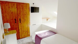 Un dormitorio con un armario de madera, una mesita de noche con lámpara, una cama con manta púrpura, un televisor montado en la pared y una ventana que deja entrar luz natural.