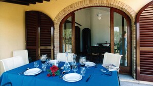 A dining table set for two with a blue tablecloth, white plates, and wine glasses, in a room with wooden shutters and a brick archway.