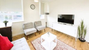 A modern living room with a white sofa, two grey armchairs, a white coffee table, a flat-screen TV on a white cabinet, and a large window with blinds. - Golden Heights 7 (Maidstone)