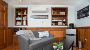 Una sala de estar con un sofá gris, estanterías de madera, una cafetera y un letrero que dice "WELCOME TO STUDIO 114".