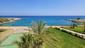 A coastal view with a tennis court, palm trees, and a clear blue sea. - Erika Apartment with Panoramic Sea View in Coralli (Paralimni)