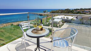 A balcony with a glass table, two chairs, and a bottle of wine. The view includes a beach and a distant town. - Erika Apartment with Panoramic Sea View in Coralli (Paralimni)