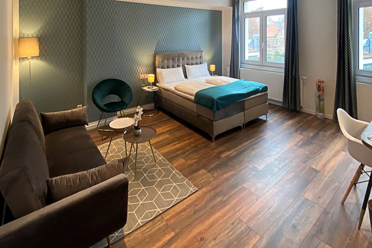 Habitación de hotel moderna con una cama grande, un área de descanso con sofá y sillón, una pequeña mesa y vistas al exterior por una ventana.