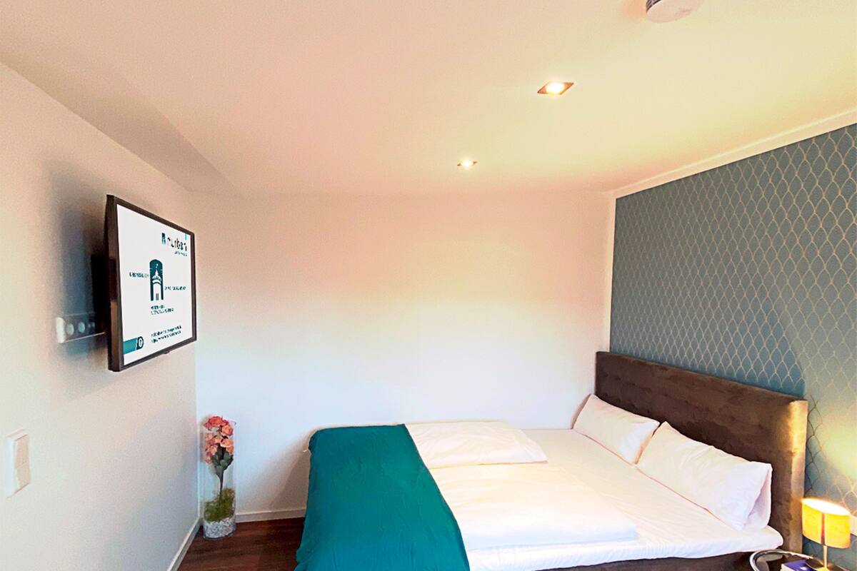 Una habitación de hotel con una cama, una mesita de noche con lámpara, un televisor montado en la pared y un cartel enmarcado.