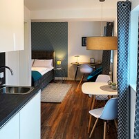 A compact hotel room with a bed, bedside table, lamp, and a small dining area with chairs and a table.