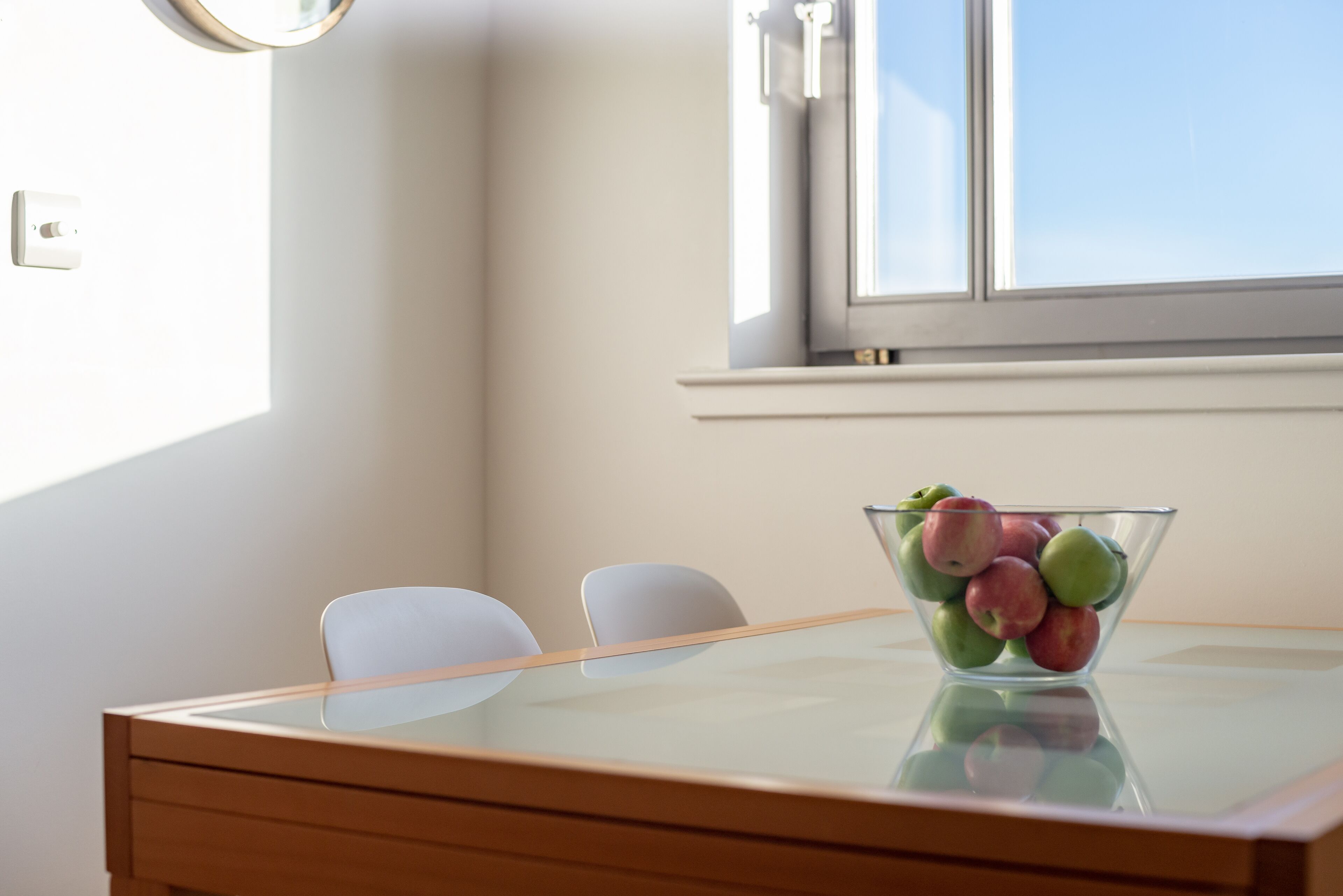 Een eettafel met een glazen schaal vol fruit, twee witte stoelen en een raam met jaloezieën.