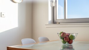 A dining table with a glass bowl of fruit, two white chairs, and a window with blinds.