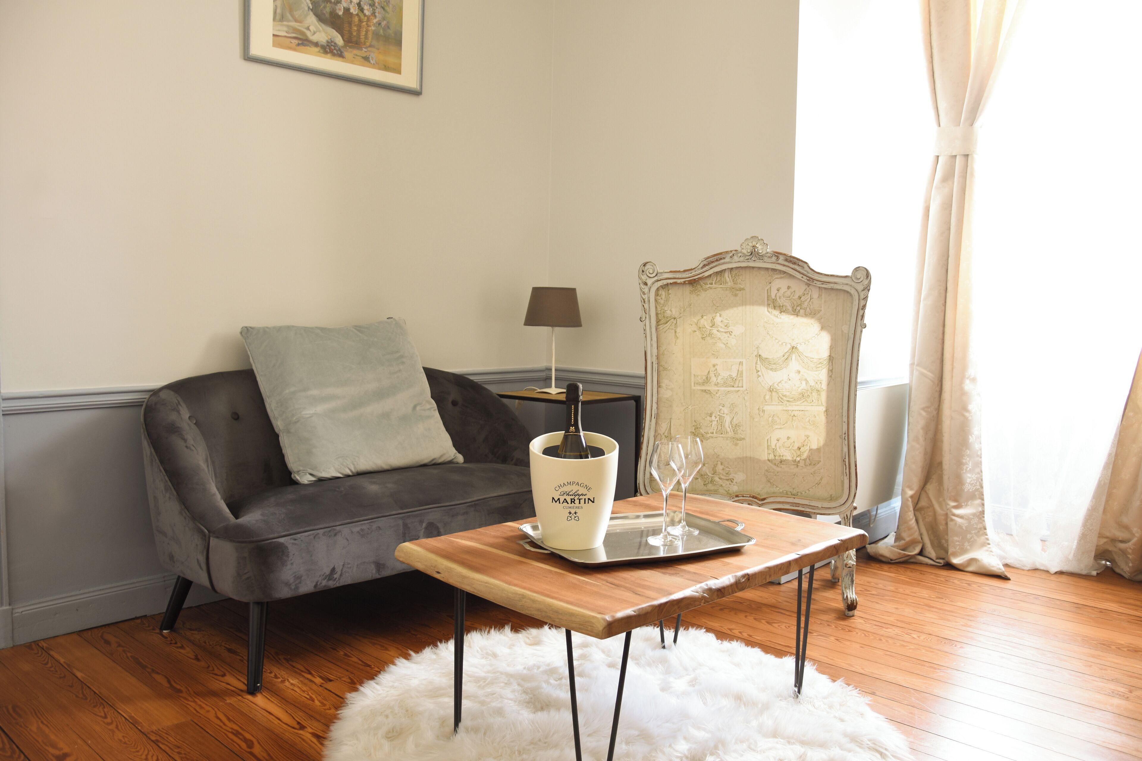 A cozy room with a grey sofa, a wooden side table with a bottle of champagne, glasses, and a tray.
