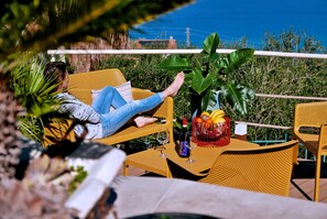 Una persona descansando en una silla amarilla con una mesita al lado, una planta en maceta y una cesta de frutas.