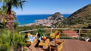 Une terrasse en bois avec des meubles d’extérieur, une vue sur une ville côtière et une montagne en arrière-plan.