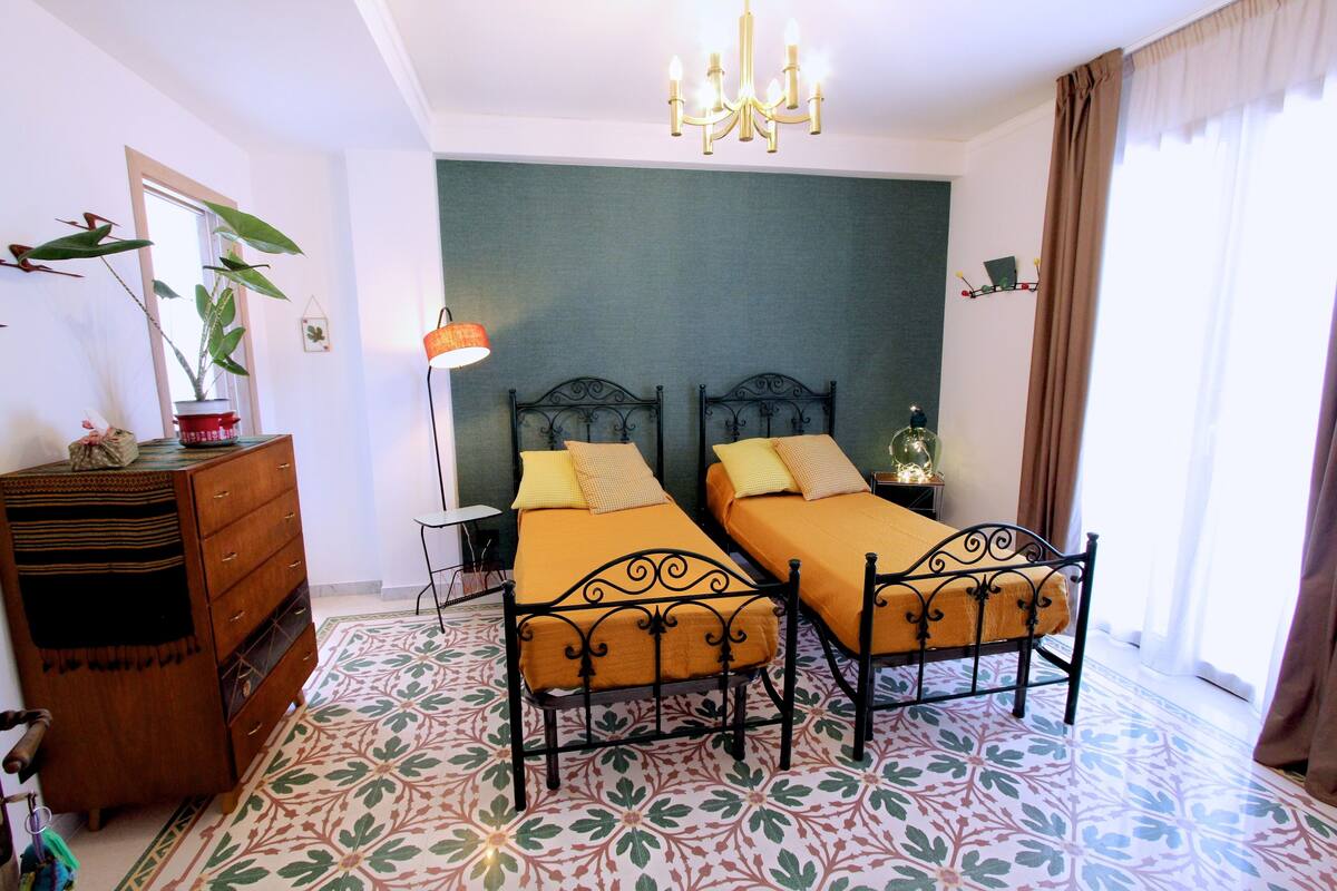 A bedroom with two beds, a dresser, a plant, and a chandelier.