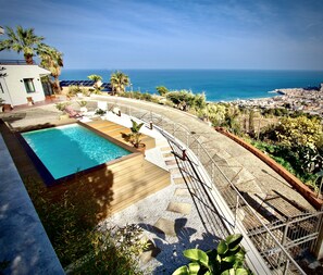 Zona de piscina con vista al océano, rodeada de follaje y una terraza pavimentada.