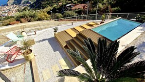 Une piscine sur le toit avec vue sur une ville côtière, une chaise rouge et une plante en pot.