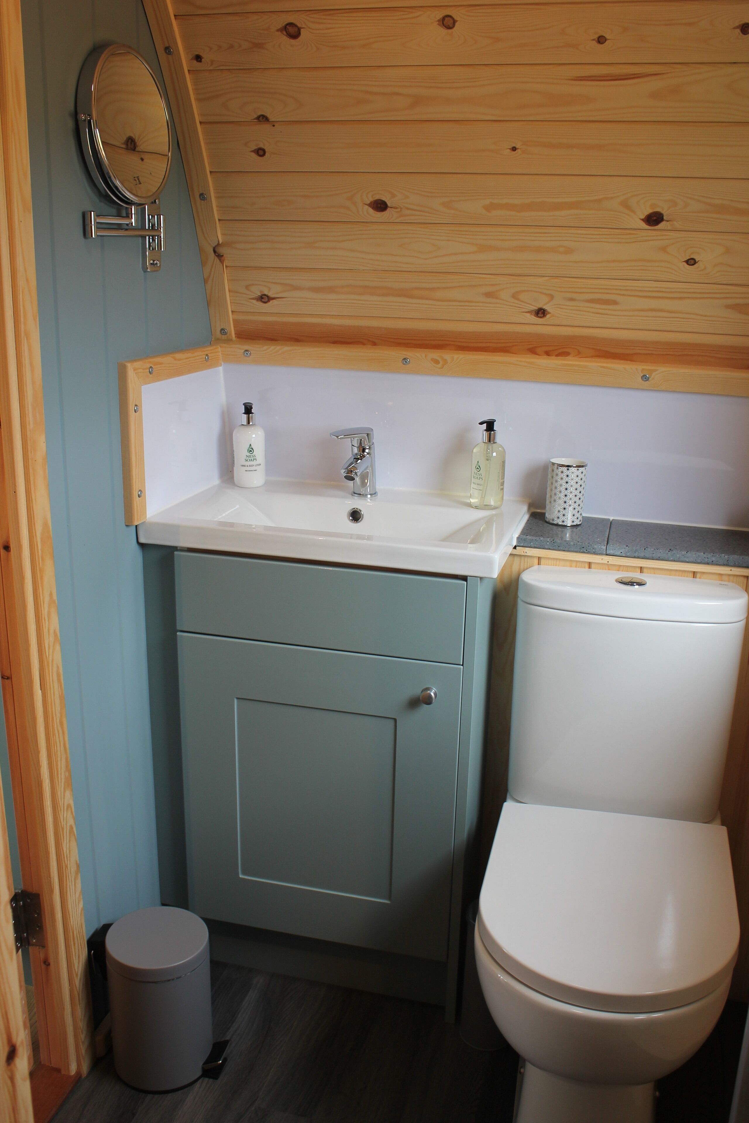 Un baño pequeño con techo de madera, un lavabo blanco con espejo, un inodoro y un cubo de basura.