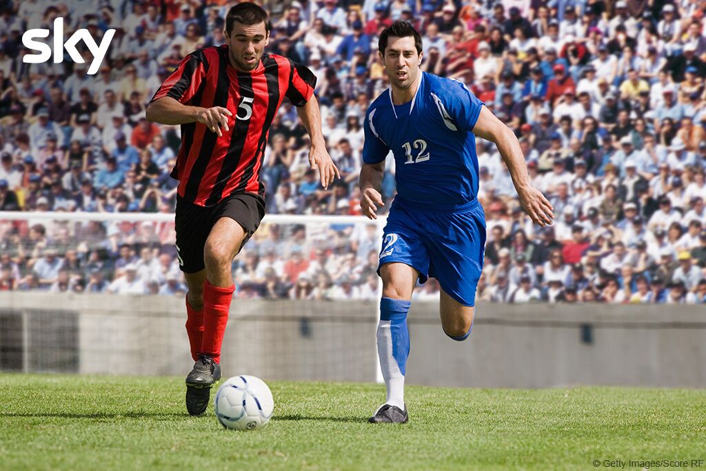 Dos futbolistas jugando en el campo, con aficionados en el fondo.