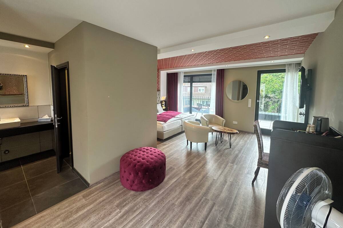 A modern hotel room with a bed, a sitting area with chairs, a small table, a large ottoman, a fan, and a TV.