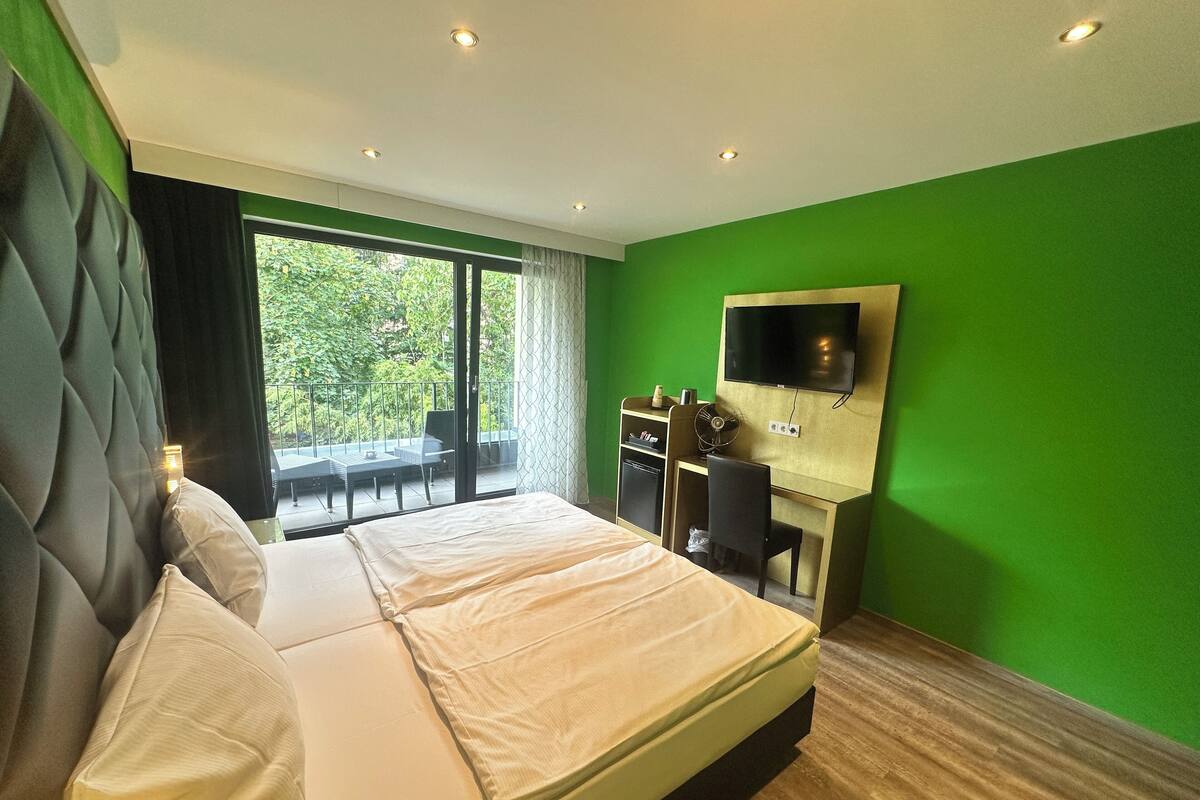 A modern hotel room with a large bed, a desk with a chair, a flat-screen TV mounted on the wall, and a balcony with outdoor seating.