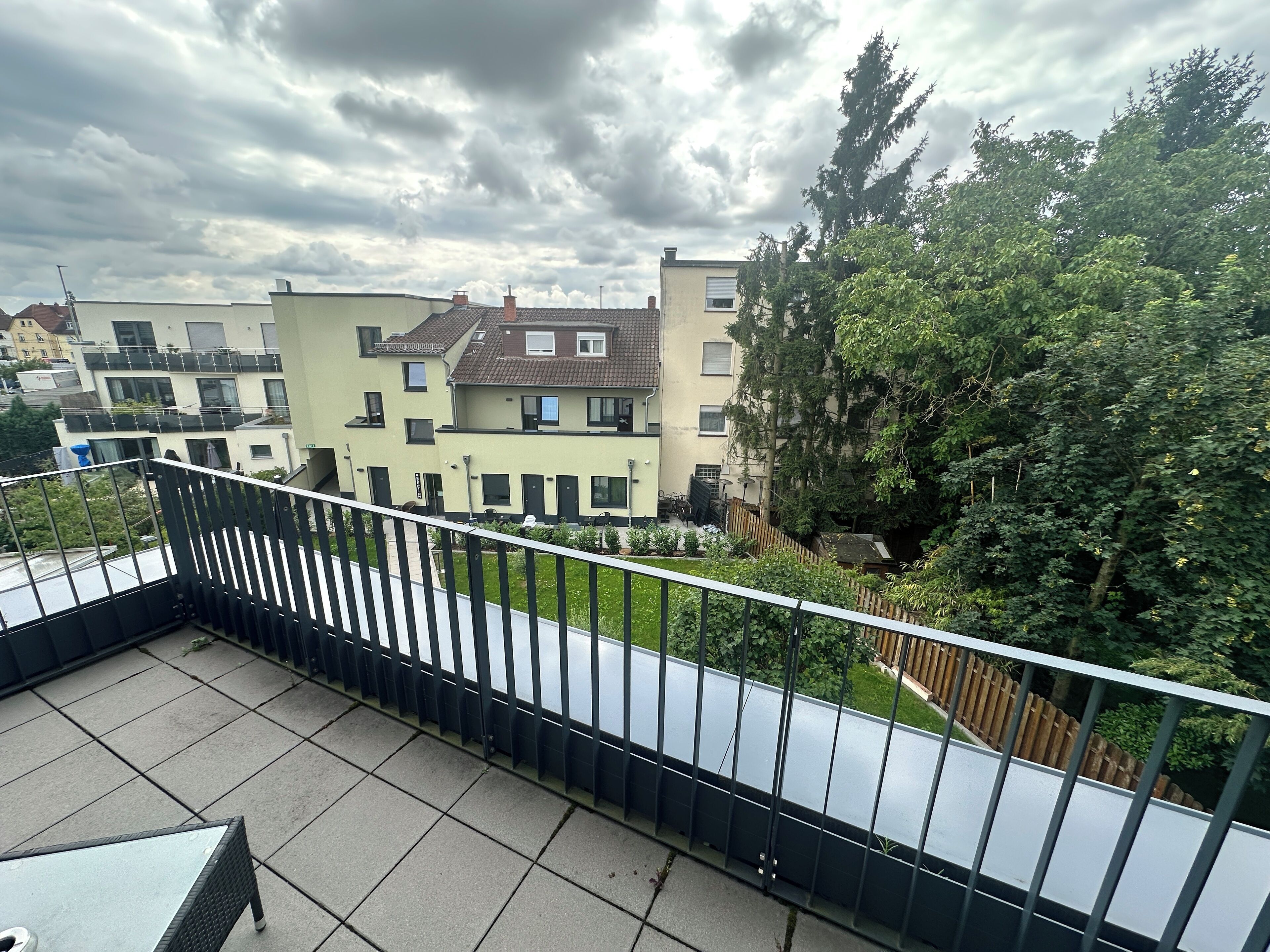 A balcony with a metal railing overlooking a residential area with multi-story buildings and a large tree.
