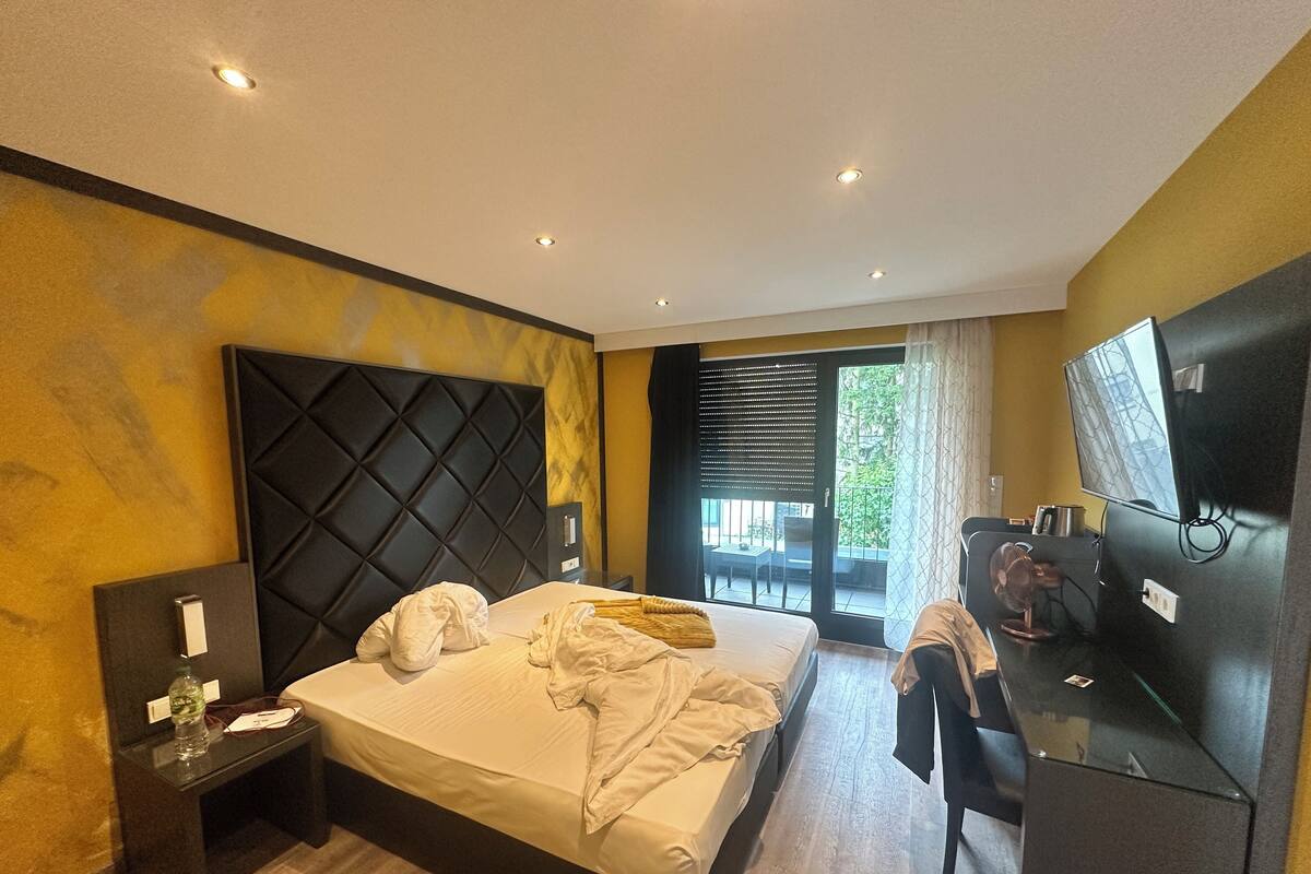 A hotel room with a large bed, a desk with a flat-screen TV, a small table with a bottle, and a sliding glass door to an outdoor area.