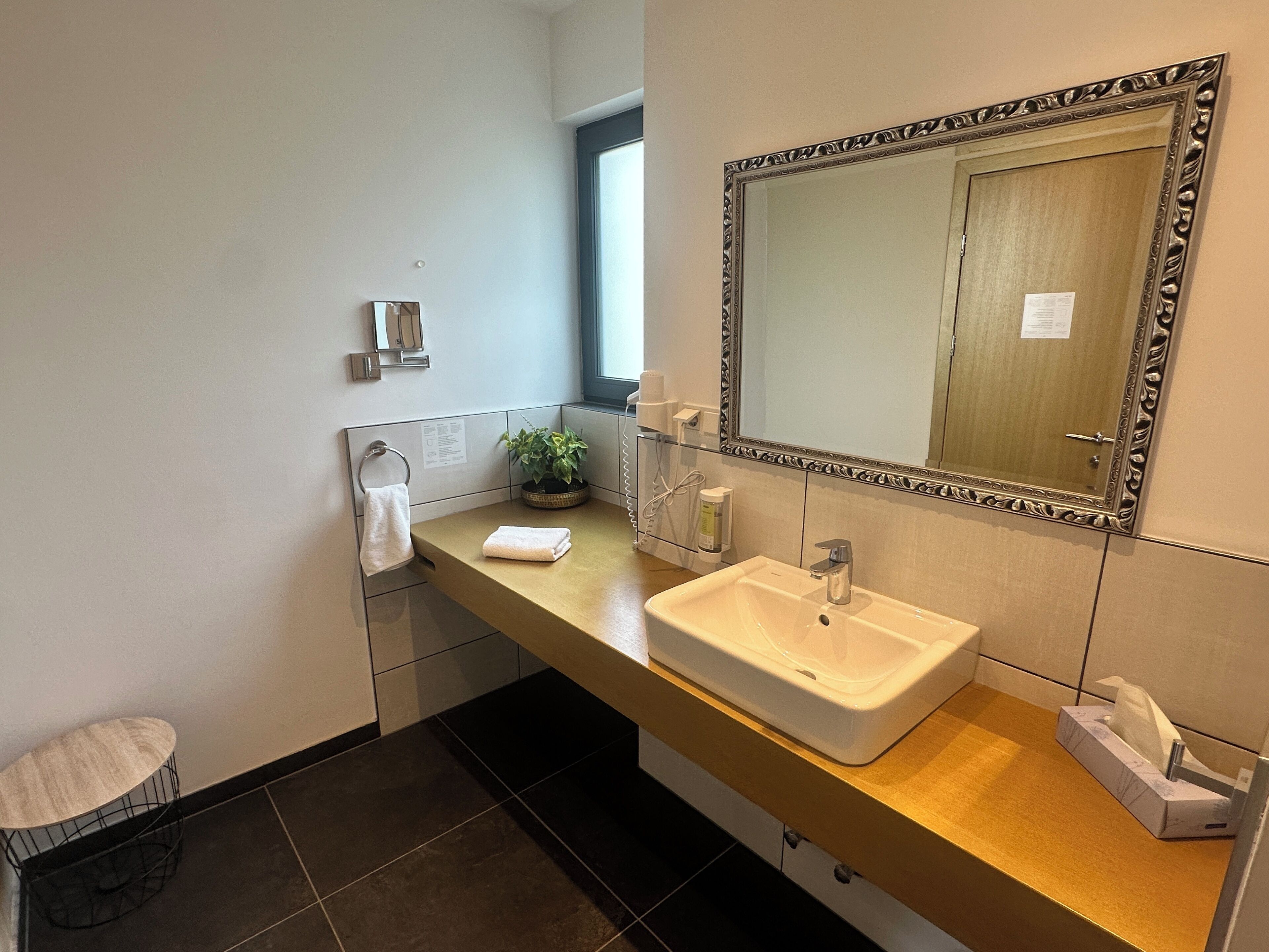 A modern bathroom with a beige countertop, a white sink, a large mirror with an ornate frame, a potted plant, and a towel rack.