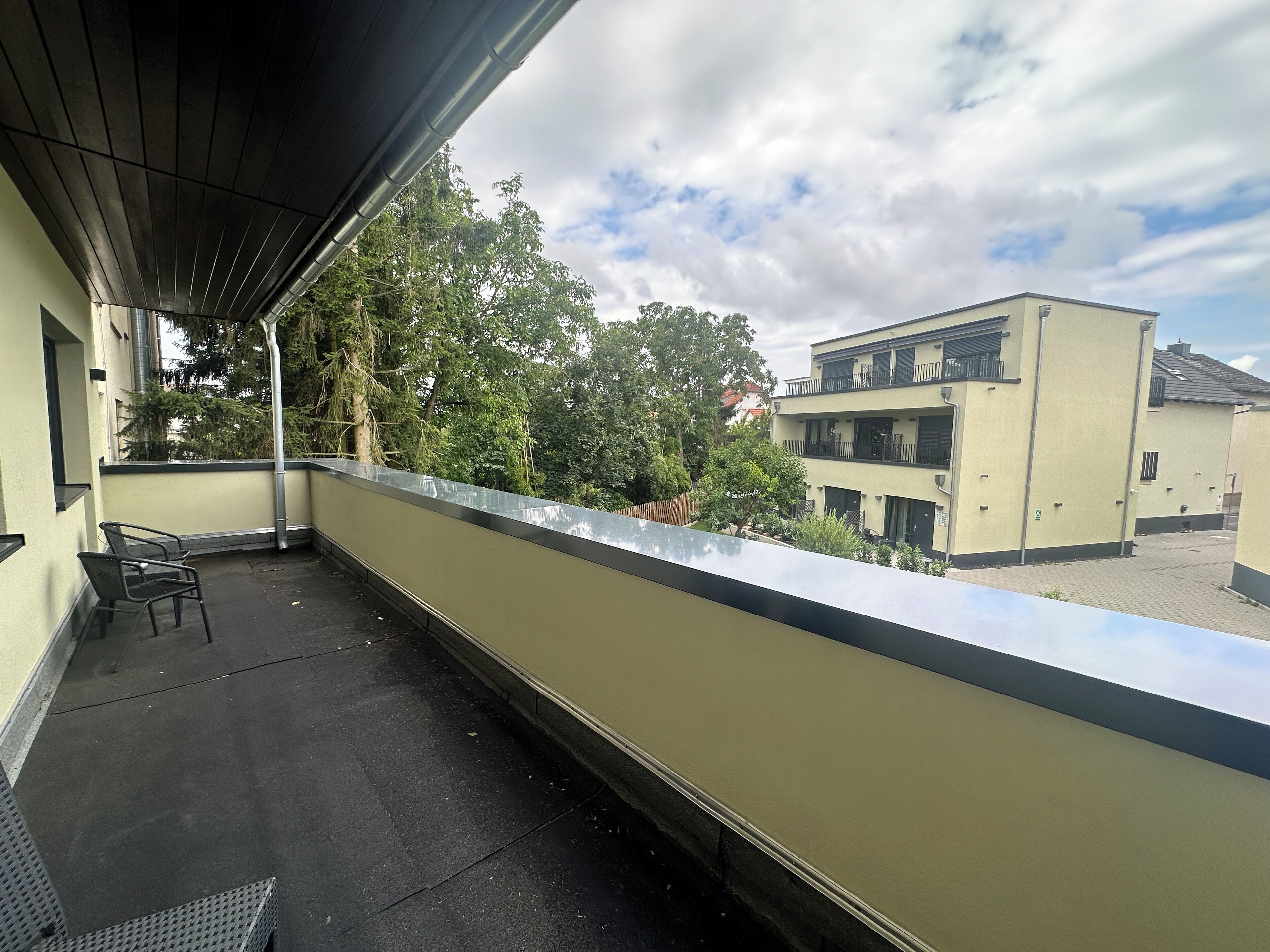 Un balcón con barandilla metálica, una silla y vista a un edificio de techo plano y múltiples ventanas.