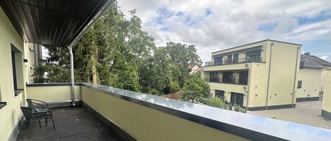 Un balcón con barandilla metálica, una silla y vista a un edificio de techo plano y múltiples ventanas.