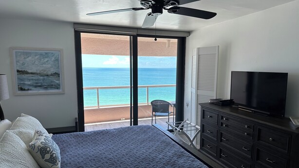 A bedroom with a bed, a TV, a ceiling fan, a painting, and a view of the ocean.