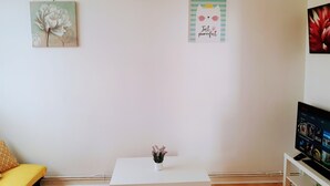 Una sala de estar con un sofá blanco, una mesita pequeña con una planta en maceta, un televisor de pantalla plana y dos obras de arte en la pared.