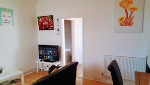 Un salón con televisor sobre un mueble blanco, un sofá negro, una mesa de comedor de madera y un radiador blanco.