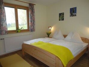 A bedroom with a wooden bed, a window with curtains, a radiator, a small plant, and two wall-mounted pictures.