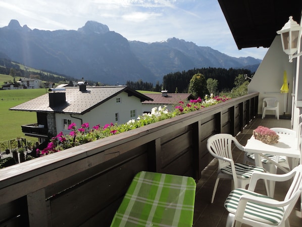 A balcony with a view of mountains, a white chair, a table, and a flower arrangement.