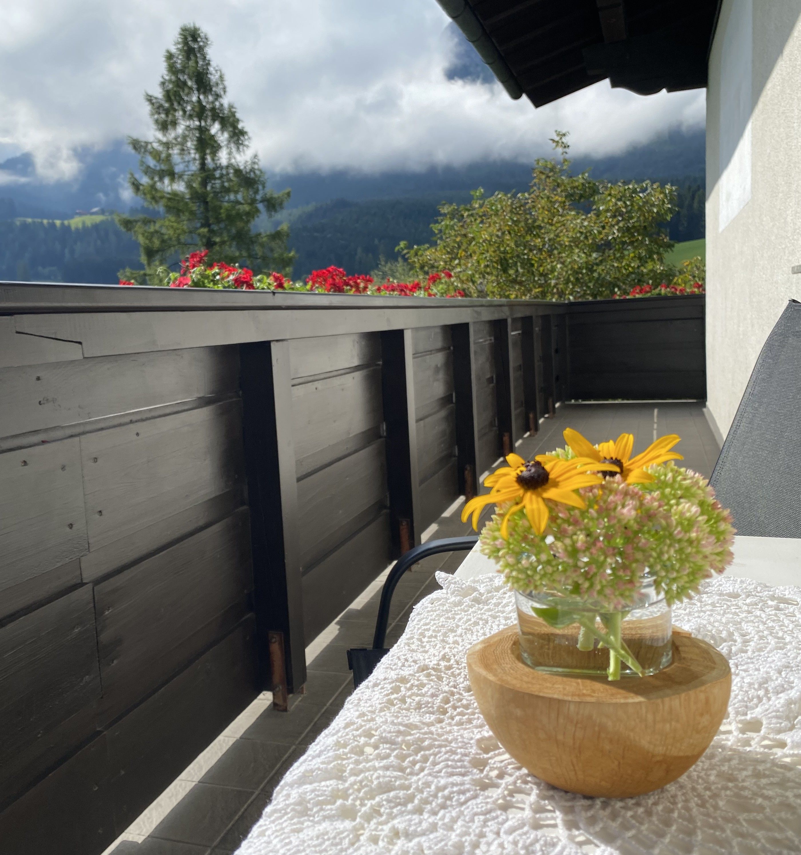 A wooden table with a white lace tablecloth, a glass vase with flowers, and a view of mountains and trees.