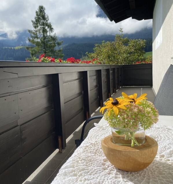 A wooden table with a white lace tablecloth, a glass vase with flowers, and a view of mountains and trees.