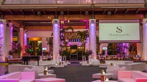 Un hall d’entrée spacieux, doté d’un grand écran affichant le logo de « Santuario », entouré de canapés et de tables blancs, et agrémenté d’une composition de plantes tropicales au centre.