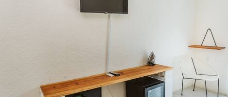 A compact room with a flat-screen TV mounted on the wall, a small wooden shelf with a remote control, a compact refrigerator, and a white chair.