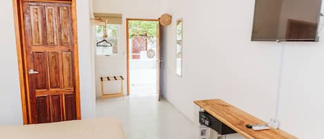 Una habitación con puerta de madera, una cama, un televisor fijado a la pared y una pequeña mesa de madera con un control remoto encima.