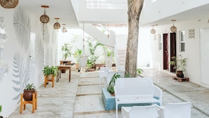 A spacious indoor courtyard with a large tree, white furniture, and hanging lights. - Ahava Hotel (Playa del Carmen)